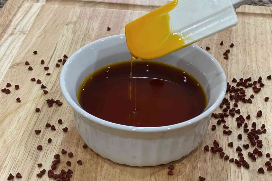 Annatto Oil in a glass bowl with annatto seeds surrounding on a wood cutting board