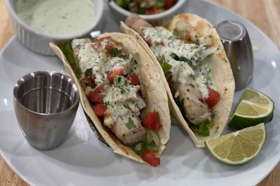 Two Hawaiian Fish tacos with lime wedges, mahi-mahi fish, creame sauce, and fresca sauce.
