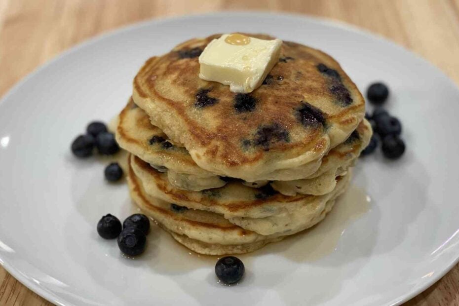 A stack of blueberry pancakes on a plate with butter and raw blueberries surrounding