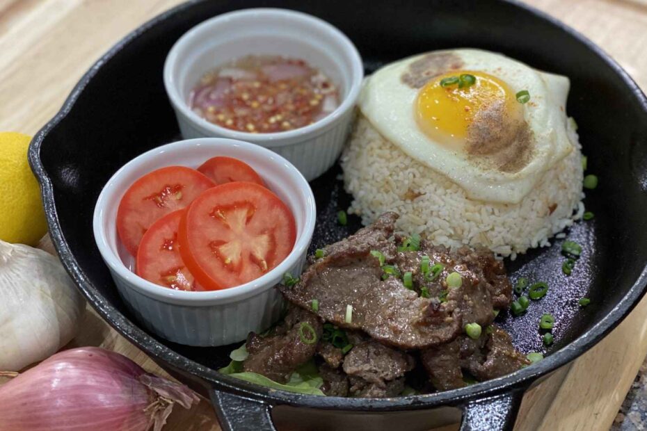 Beef Tapa prepared in an iron skillet with a side of rice with an egg over the top